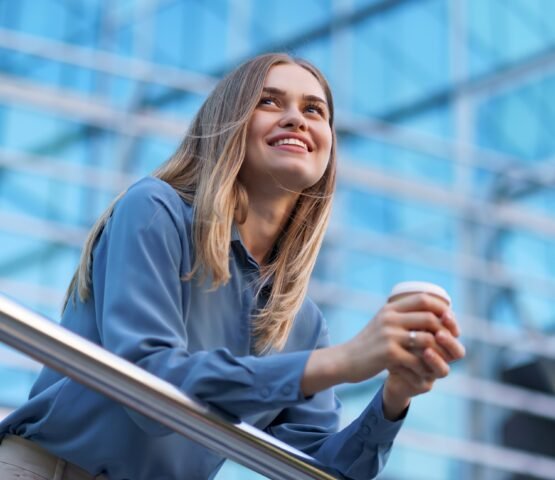 Young smiling professional woman having a coffee break during her full working day. She holds a paper cup outdoors near the business building while relaxing and enjoying her beverage.